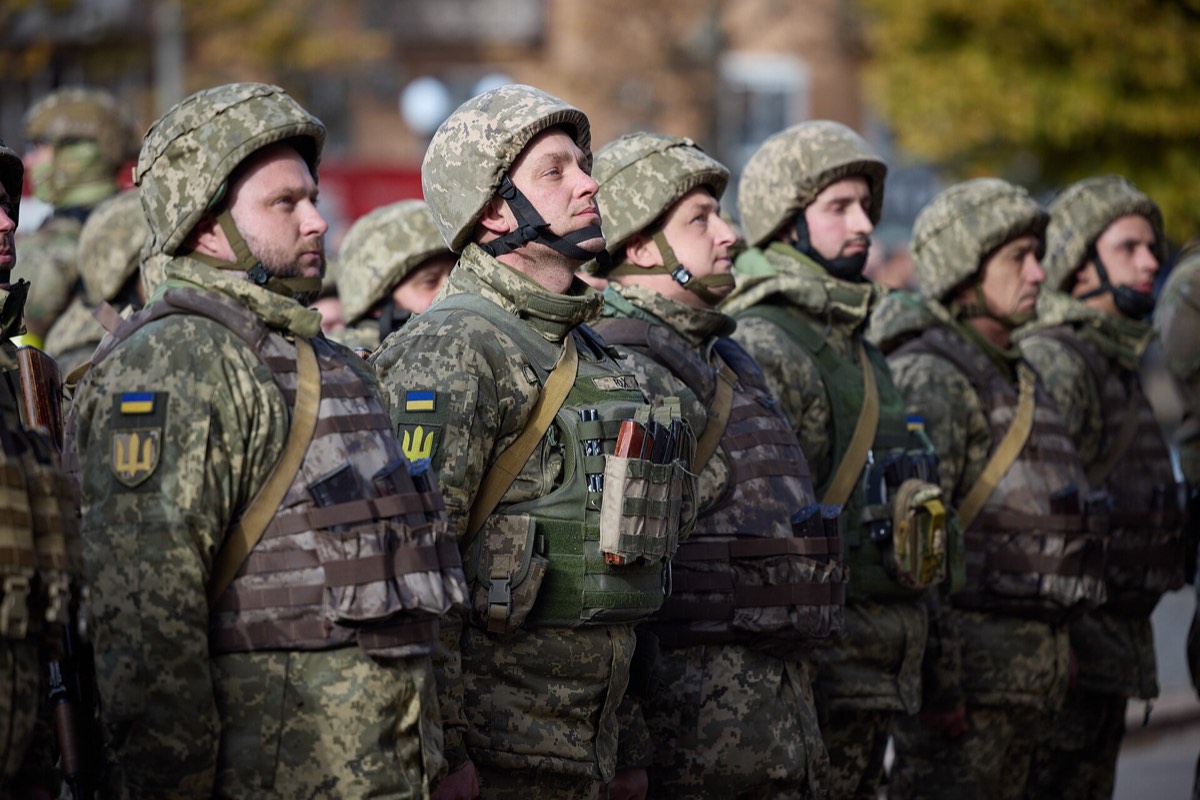 Ukrainian soldiers standing in formation in Kherson, in helmets and body armour with Ukrainian flag and trident patches, looking up at something above the frame.